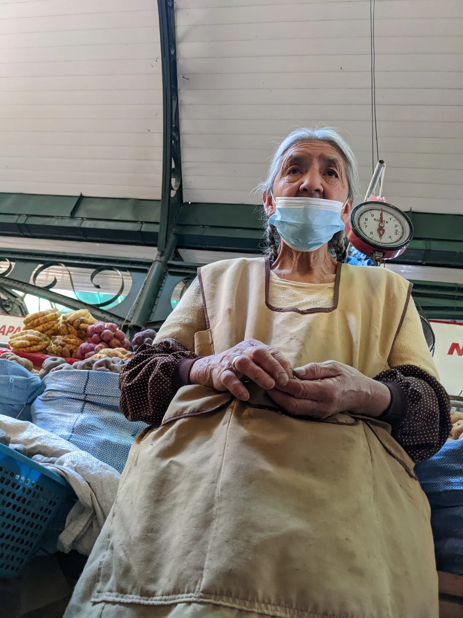Elderly woman vendor wearing a blue medical mask at a market stall in Cochabamba.