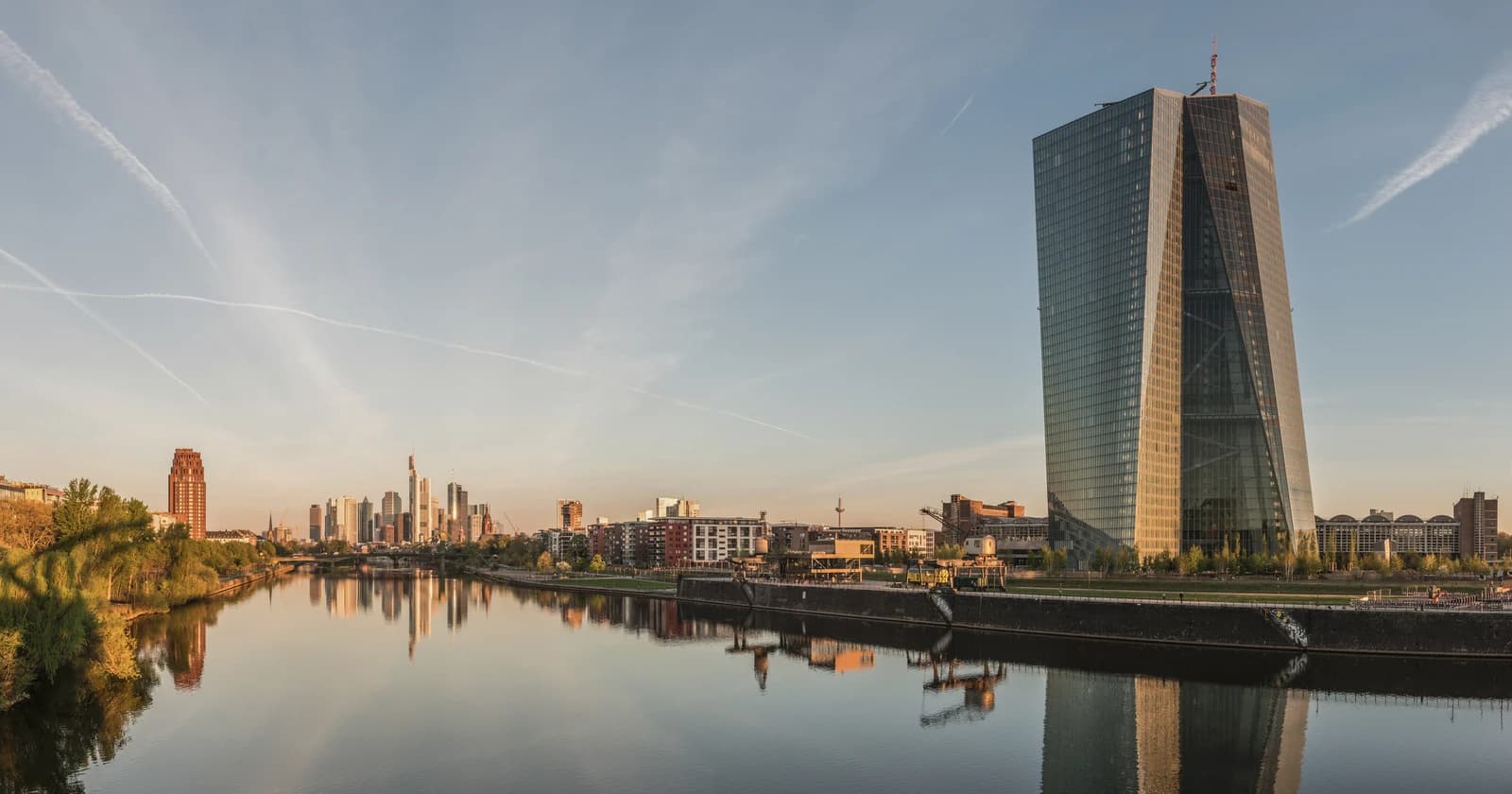 The European Central Bank building stands prominently alongside the Frankfurt skyline reflected in a river at dawn.