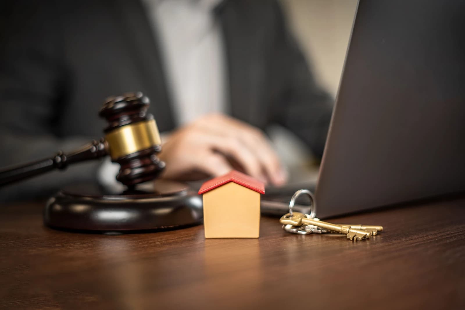 A gavel, miniature house, and keys sit on a desk next to a person using a laptop.