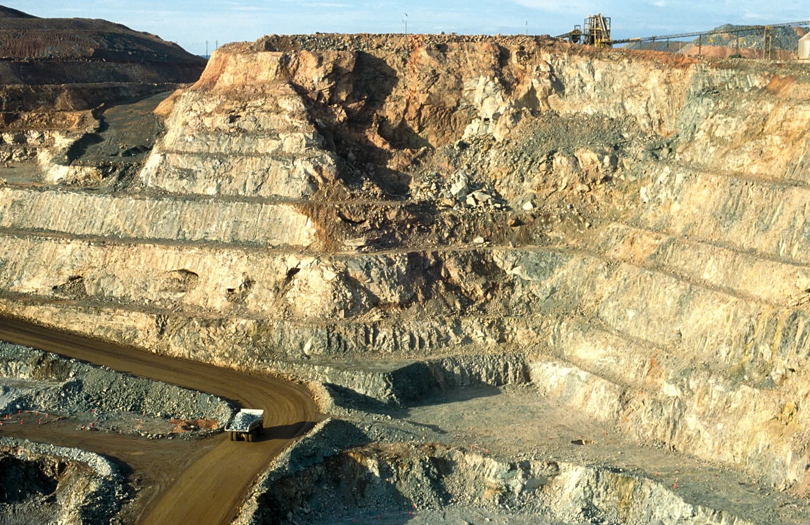 A large ore truck travels on a dirt road within an open-pit gold mine.
