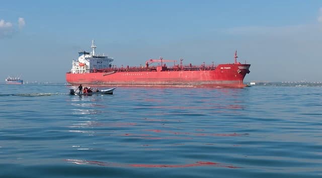 A large red oil tanker cruises on blue water with a small fishing boat nearby.