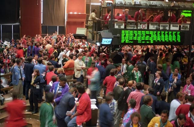 Traders in a busy, crowded trading pit, looking at screens and communicating.