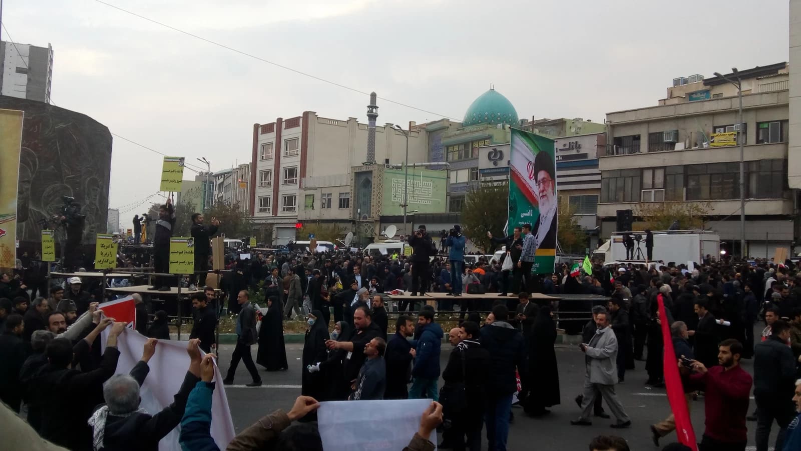 Large crowd of Iranians gathered in a city square with banners and a large portrait of a leader.