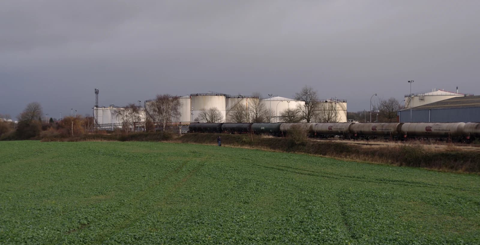 Freight train with multiple tank cars on railway tracks next to large industrial storage tanks.