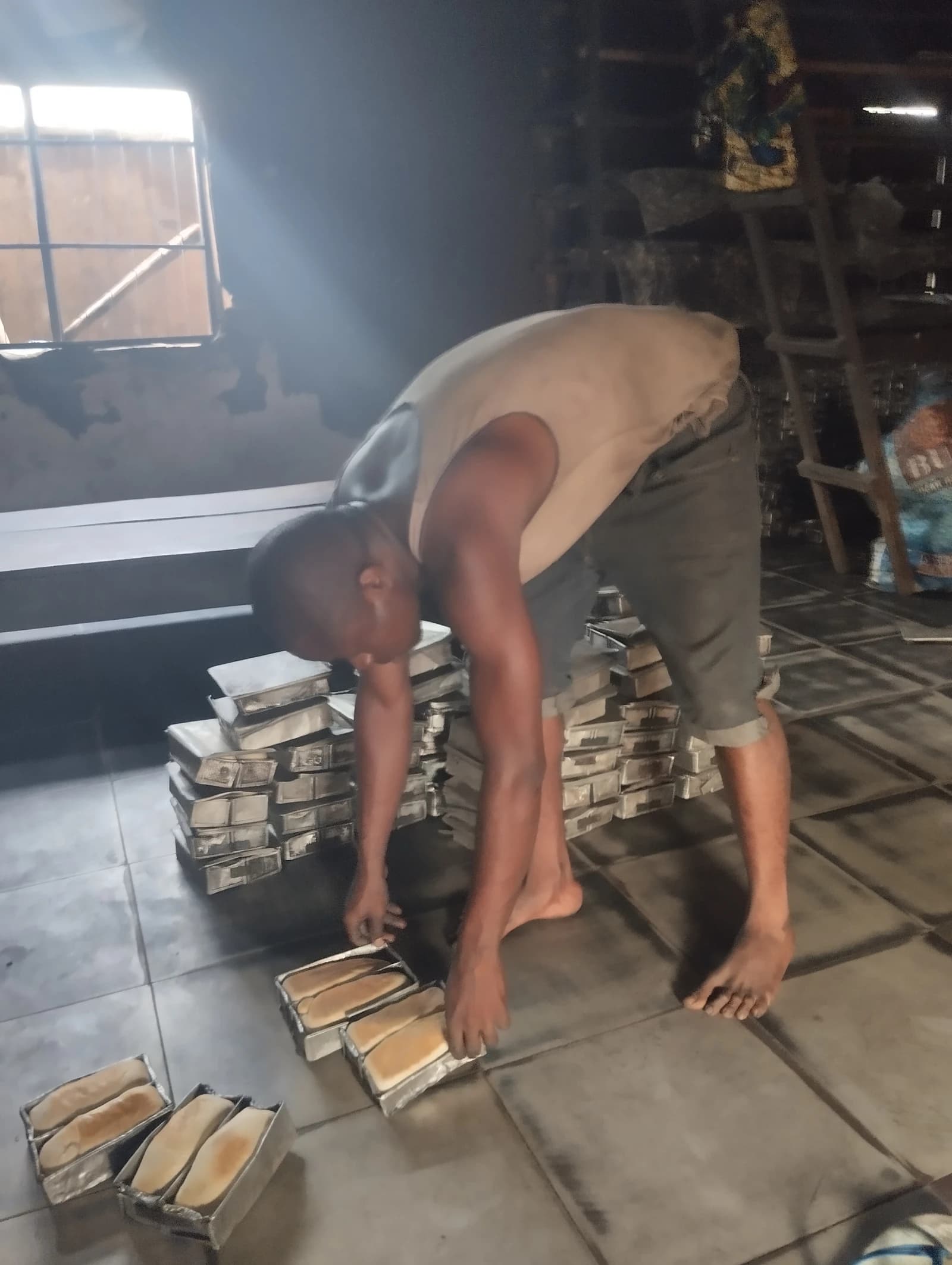 A person bending over, arranging freshly baked bread loaves in metal pans on the floor.