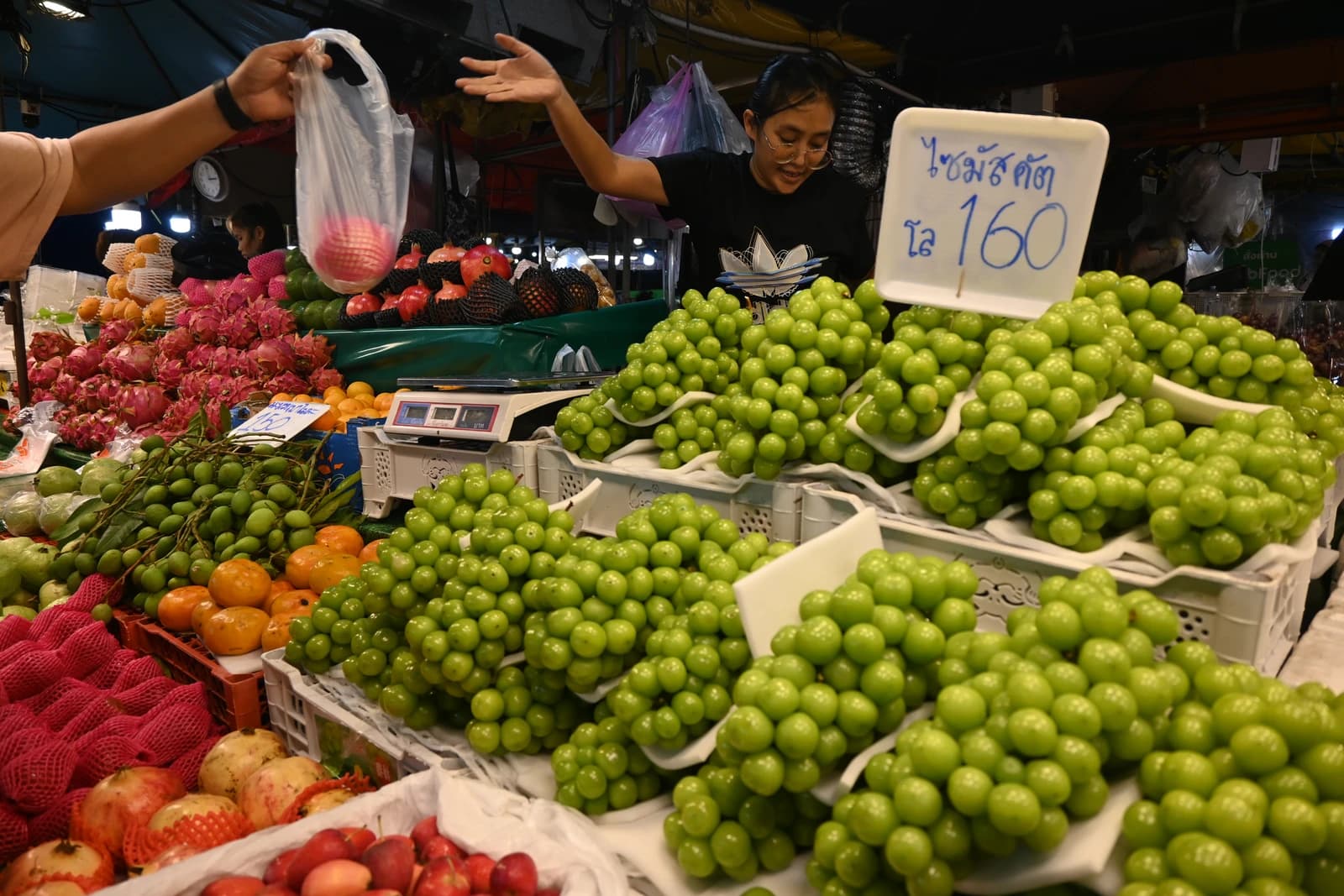 Vendor hands a bag to a customer at a market stall piled high with grapes.