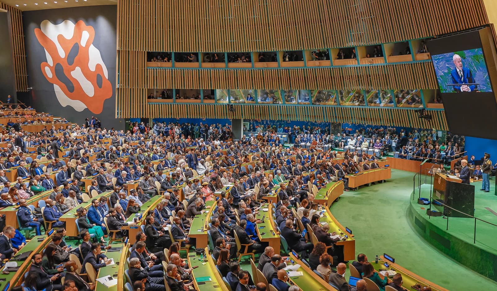 A wide shot of a large assembly hall filled with delegates, with Brazil's President at a podium speaking to the group, and his image shown on a large screen above.