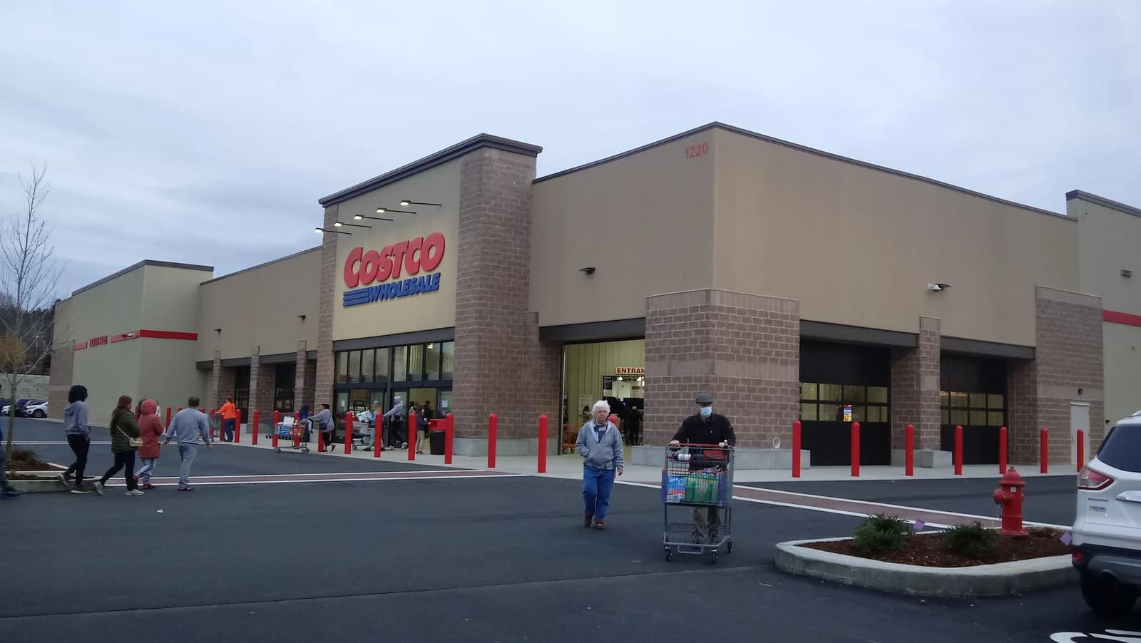 People walking outside a Costco Wholesale Club store, one person pushing a shopping cart.