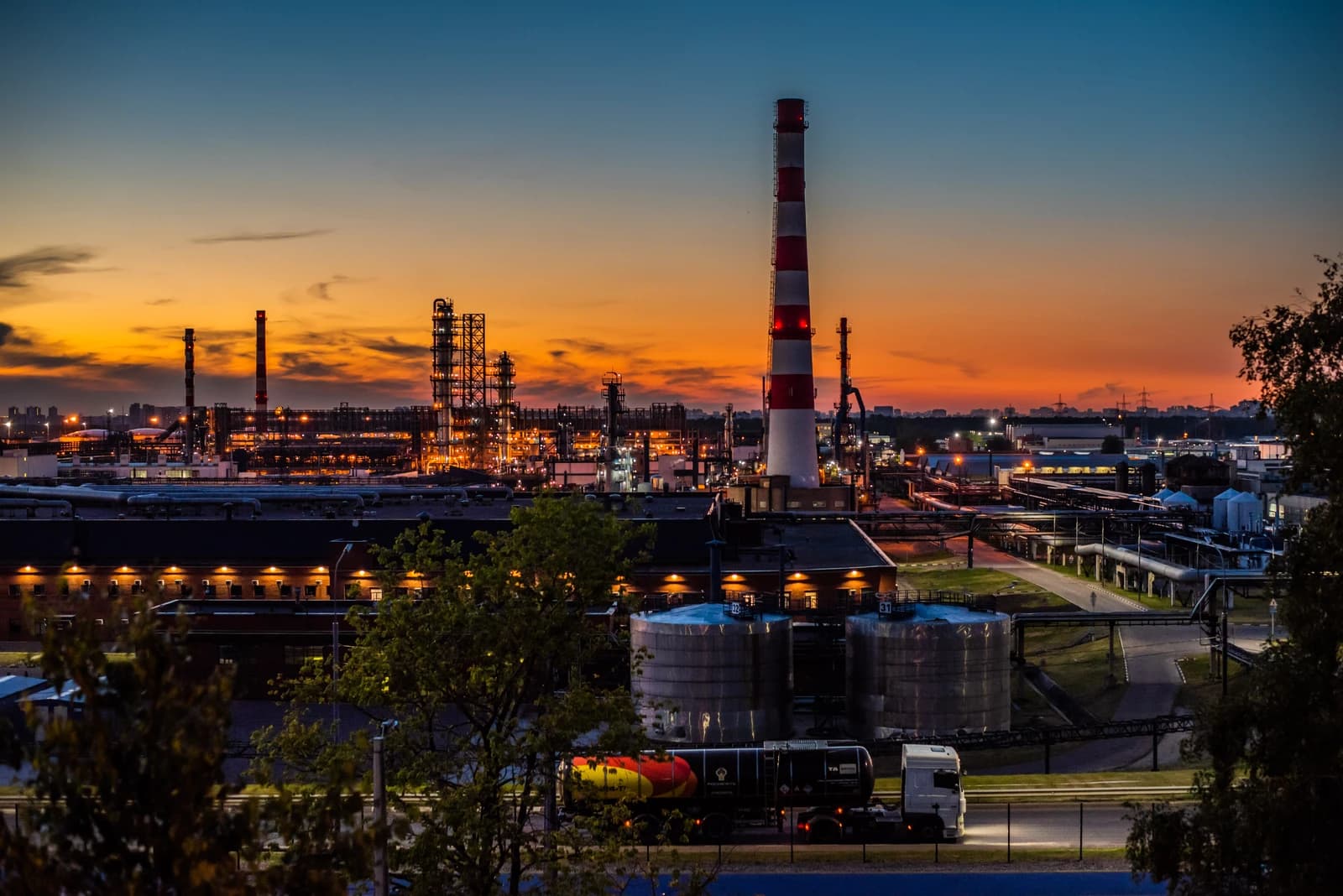 An illuminated oil refinery in Moscow with storage tanks and a large smokestack at dusk.