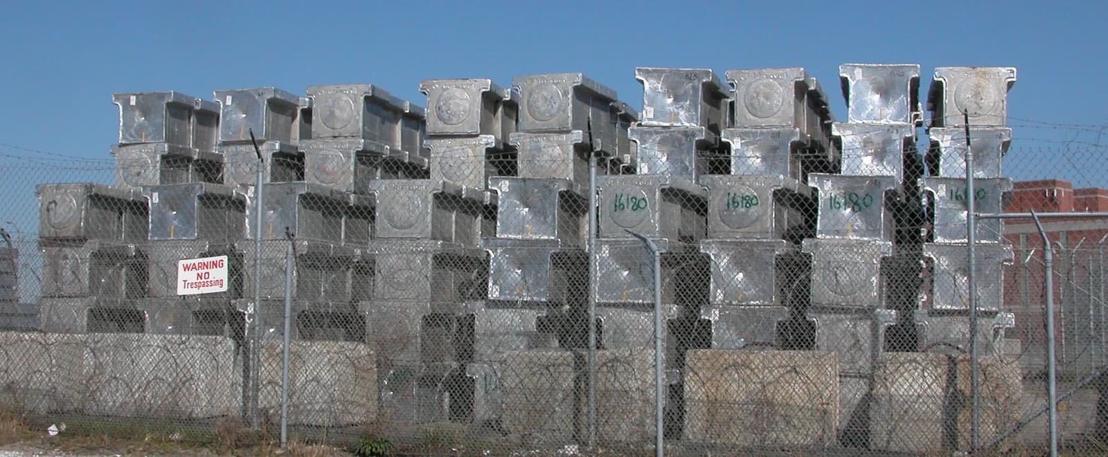 Stacks of large, grey aluminium ingots visible behind a chain-link fence.