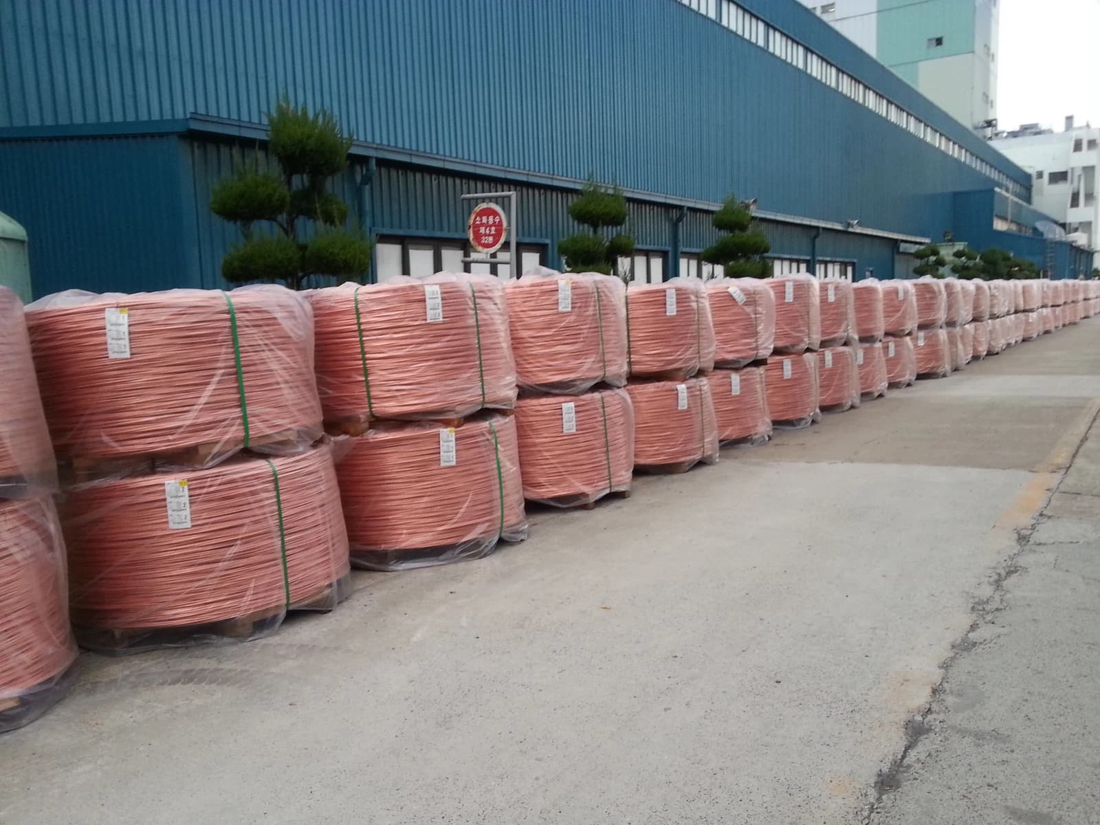 Bundles of copper wire rod coils stacked outdoors next to a factory building.