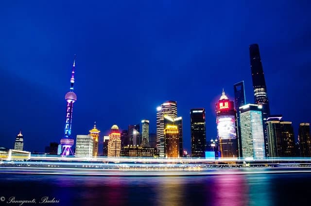Skyscrapers of Shanghai's Pudong Financial District illuminated at night with light trails on the water.