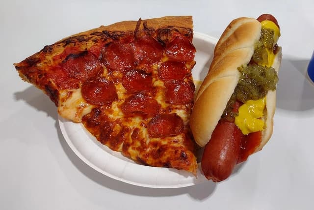 A slice of pepperoni pizza and a hot dog with mustard and relish on a white paper plate.