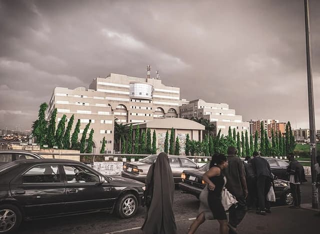 Traffic and pedestrians on a street in Abuja, Nigeria, with a large building in the background.