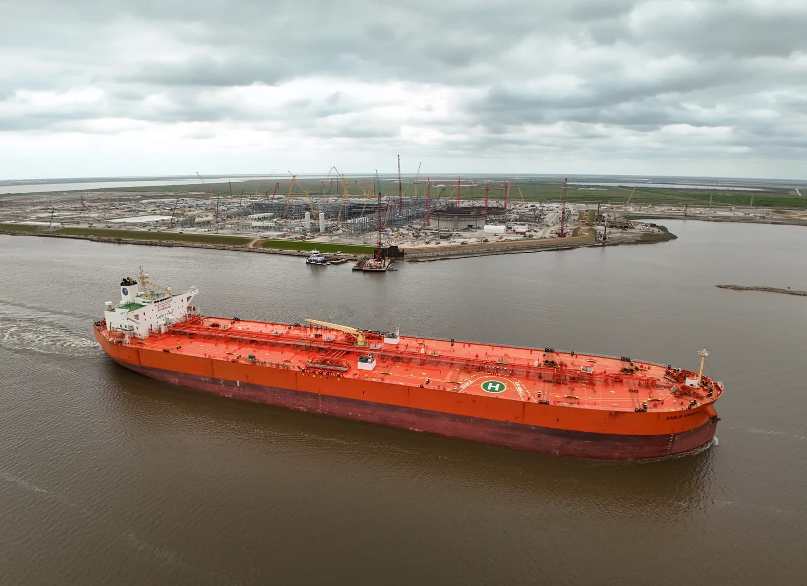 An orange crude oil tanker moves along a waterway past an industrial complex.