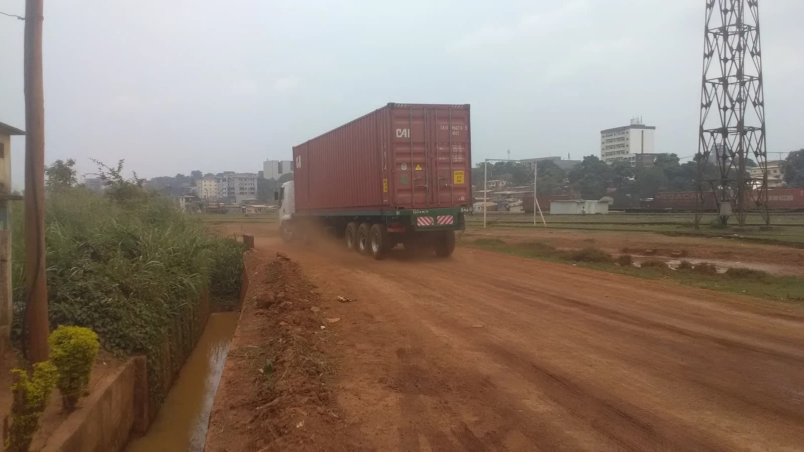Semi truck carrying a red shipping container on a dirt road with buildings in the background.