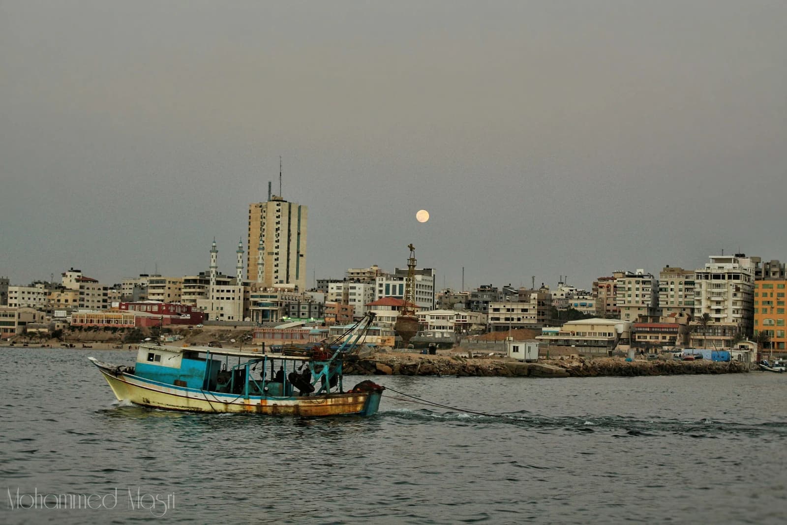 Fishing boat sails on water with the skyline of Gaza city in the background.