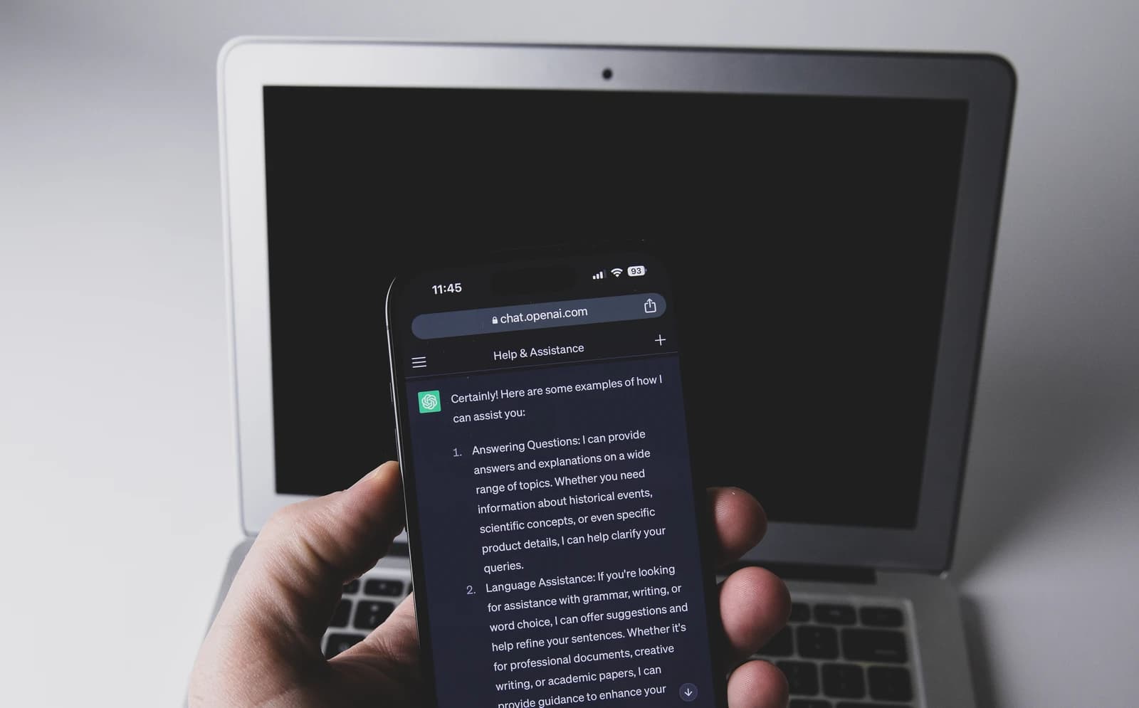 Hand holding a smartphone displaying the ChatGPT app, a laptop is in the background.