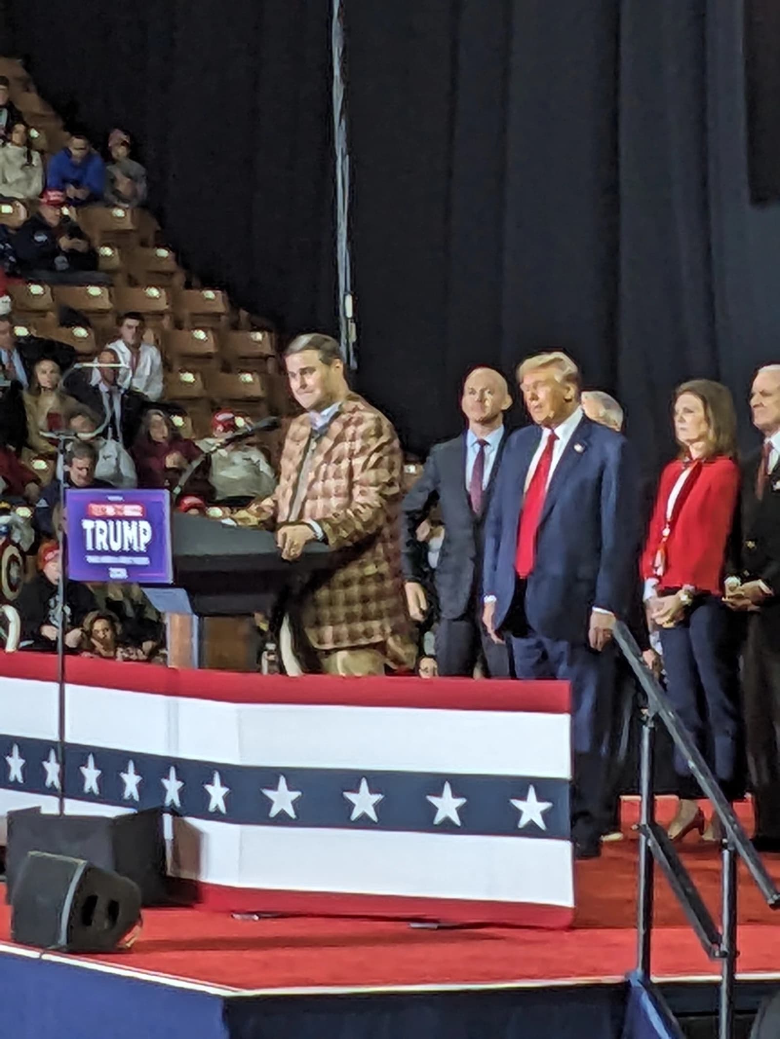 Donald Trump, former President, standing on a stage at a rally with other individuals.