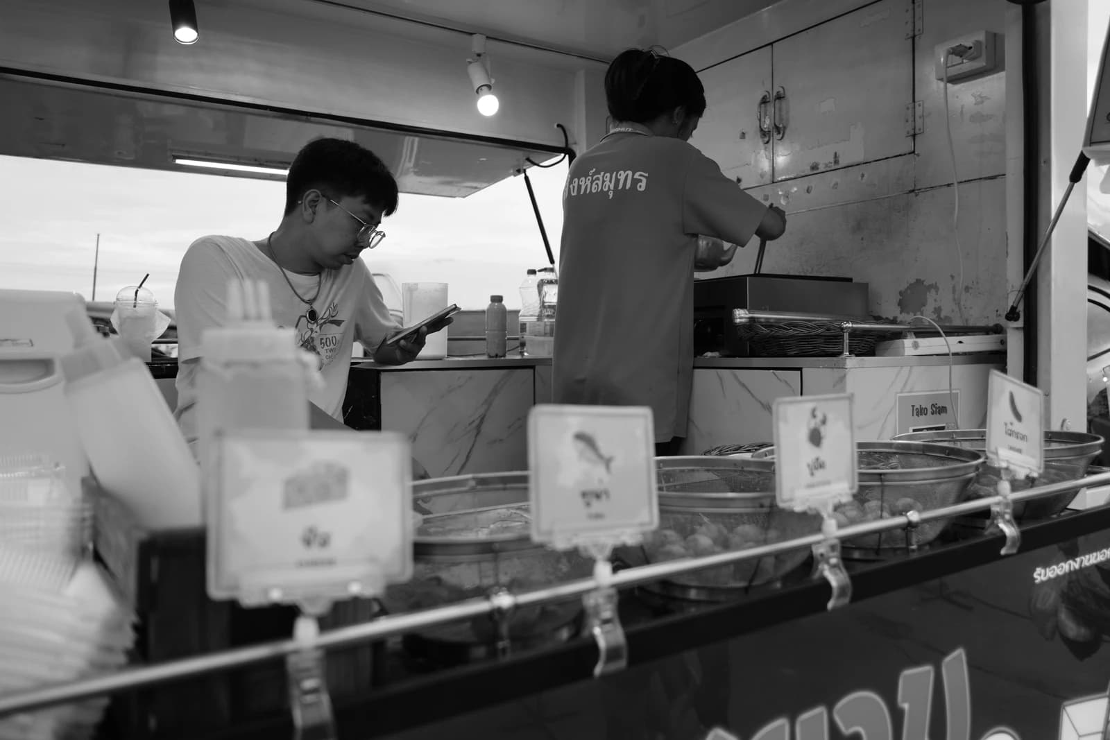 Two street food vendors work behind a counter, one prepares food, the other checks a phone.