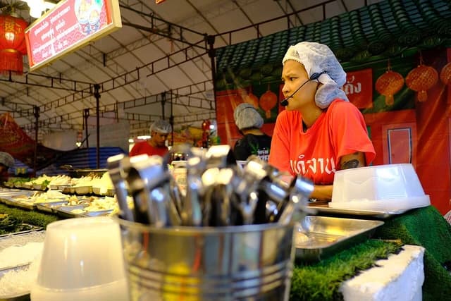 Vendor wearing a hairnet and headset stands behind a brightly lit food stall.