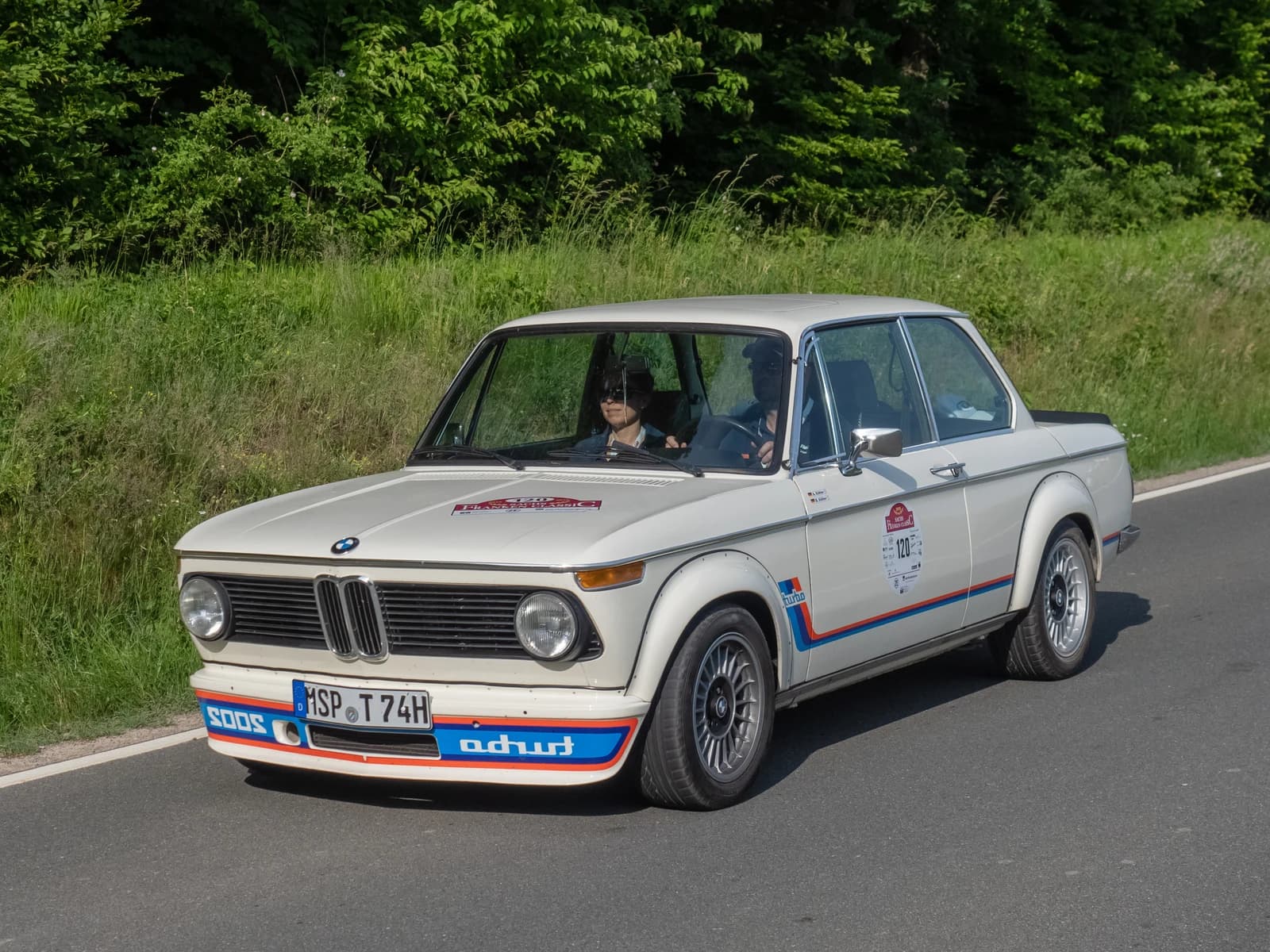 White BMW 2002 Turbo classic car with a driver and passenger on a road.