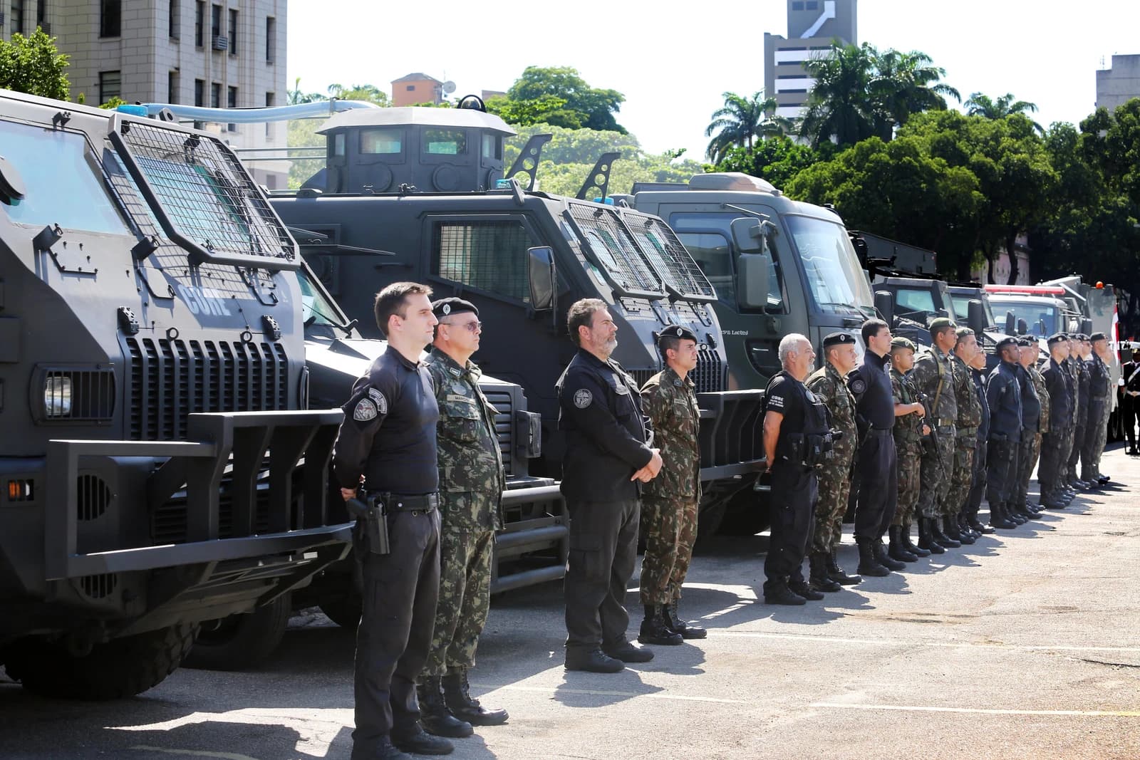 Military and police personnel stand in a line next to several armored vehicles outdoors.