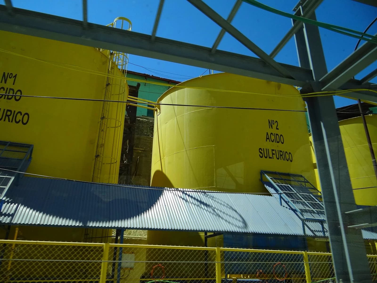 Large yellow tanks labeled 'Acido Sulfurico' in an industrial area under a blue sky.