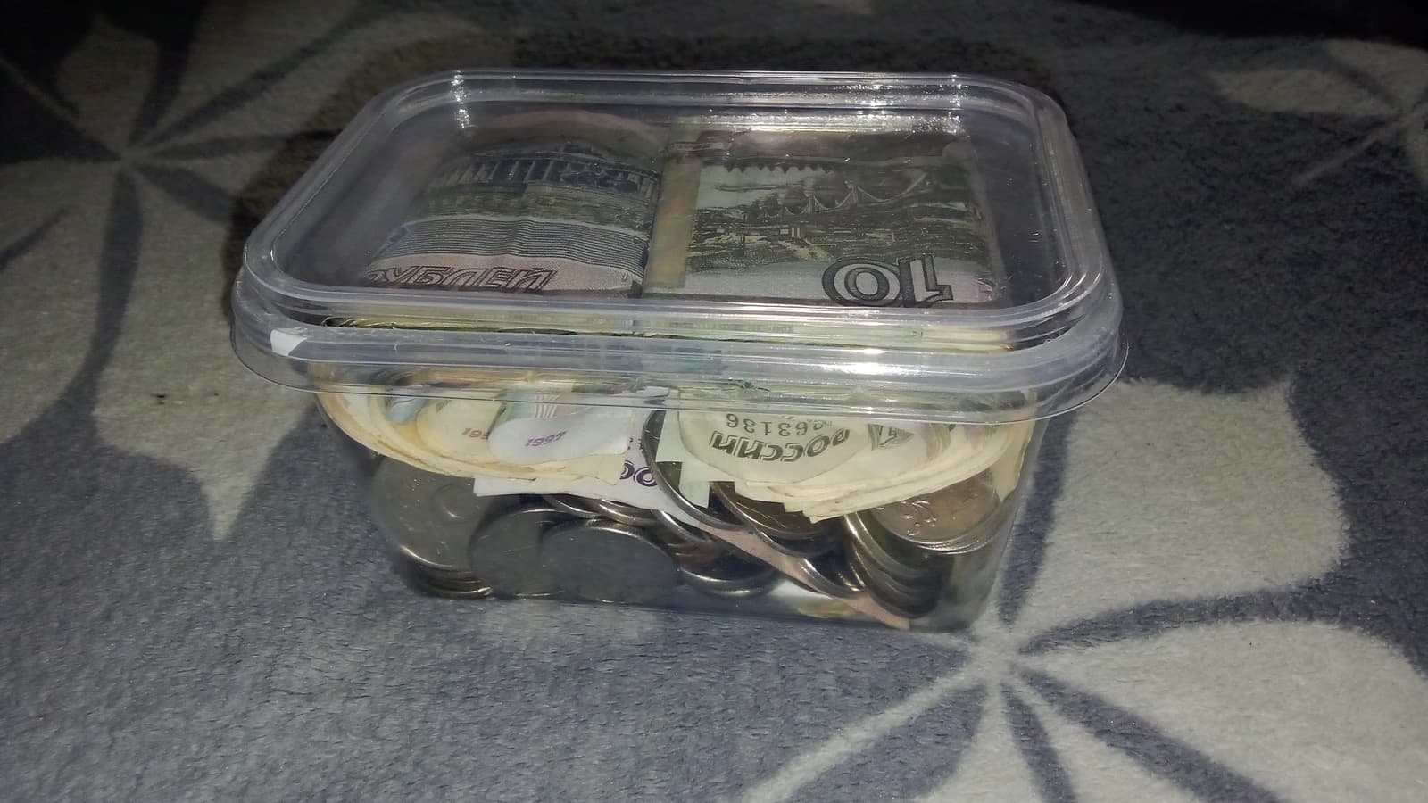 A clear plastic container, functioning as a piggy bank, filled with various coins and banknotes.
