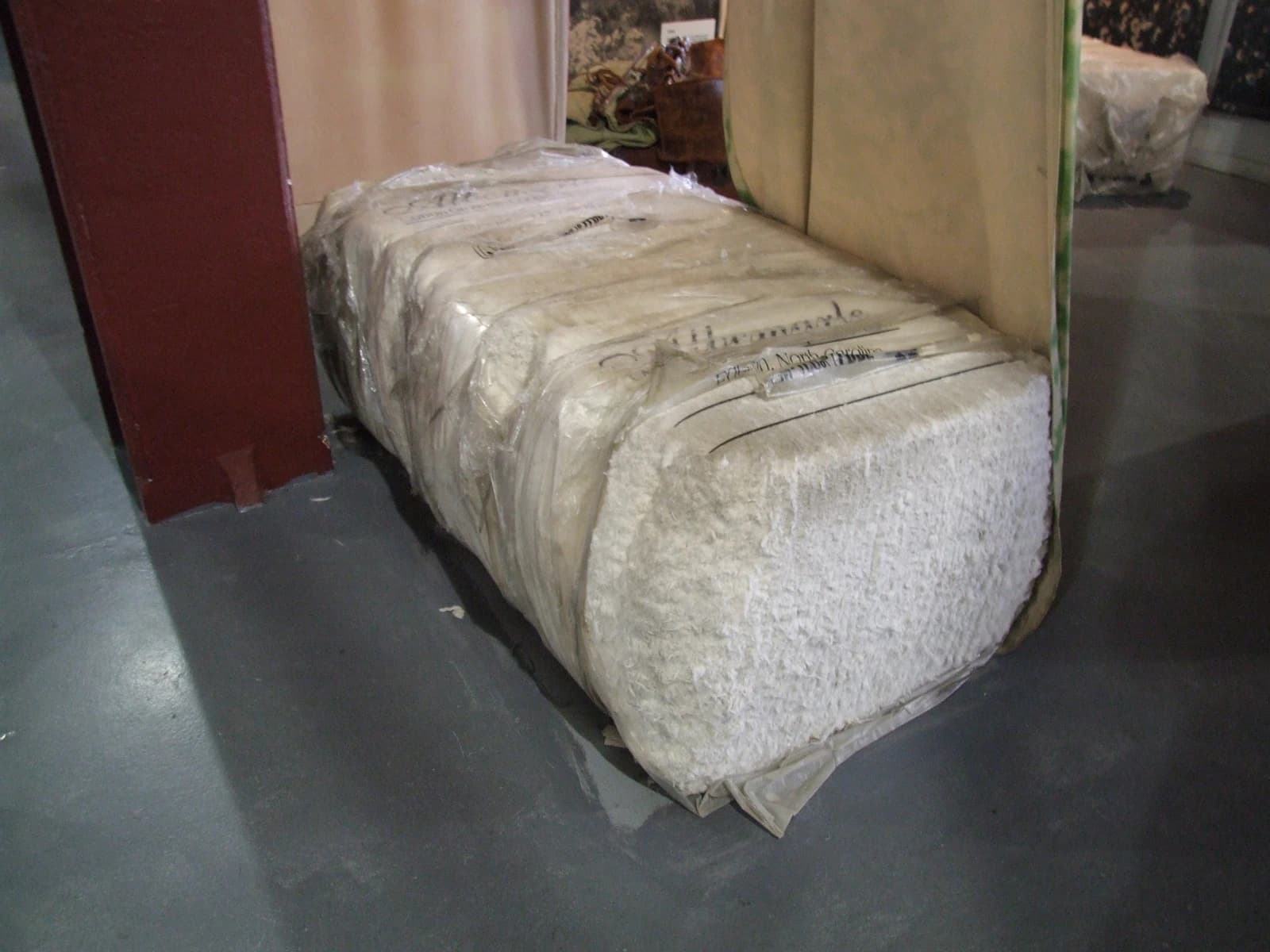 A white bale of raw cotton wrapped in clear plastic on a dark floor.