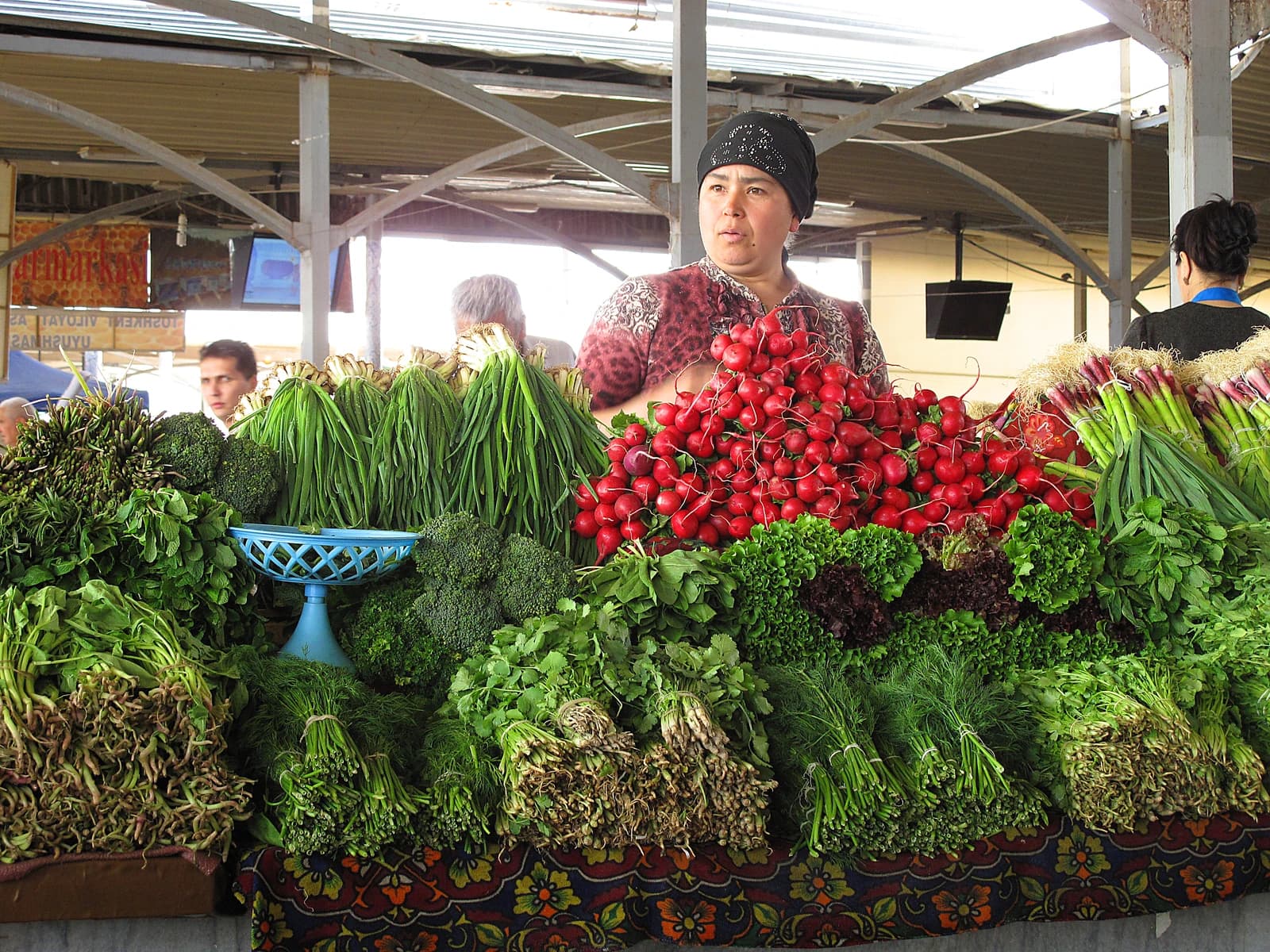Vendor stands behind a colorful display of fresh radishes, green onions, and other vegetables.